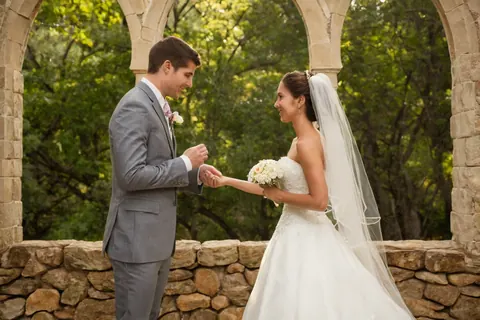 A bride and groom exchange wedding rings under stone arches in a forest setting.