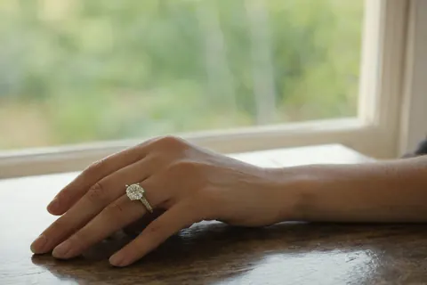 A hand wearing a diamond engagement ring on a wooden table near a window