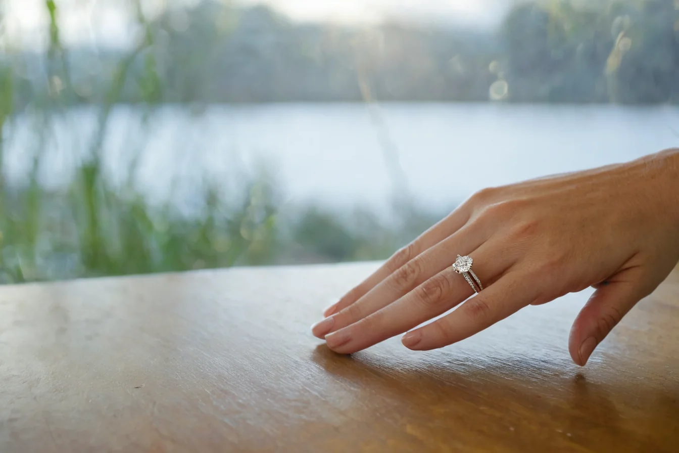 A hand wearing an engagement ring on a wooden table near a lake