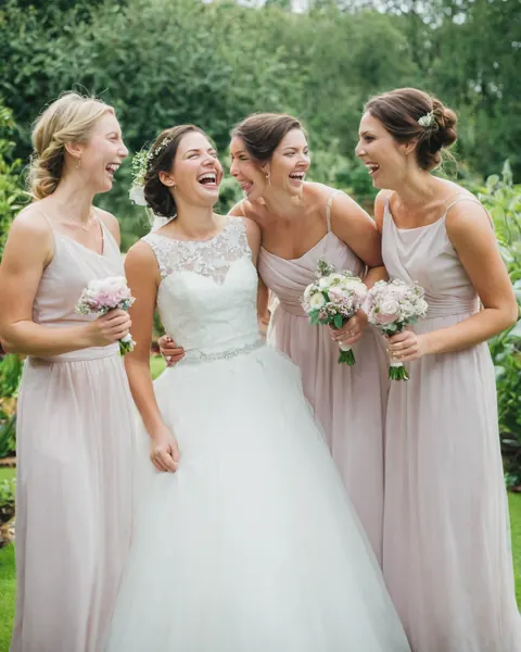 Four women in wedding attire laughing together in a garden holding bouquets