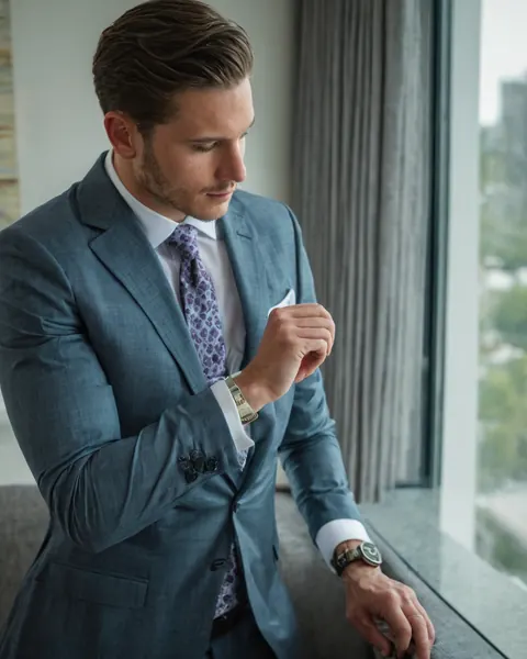 Man in blue suit checking watch by window with city view