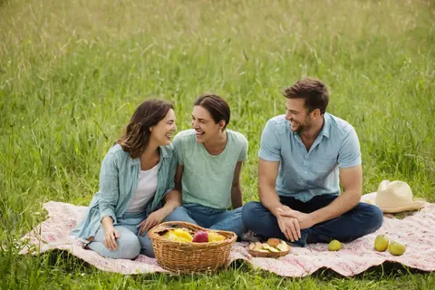 Three people laughing during a picnic in a grassy field with a basket and fruit.