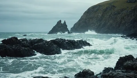 Waves crash against dark rocks near a steep cliff under a gray sky