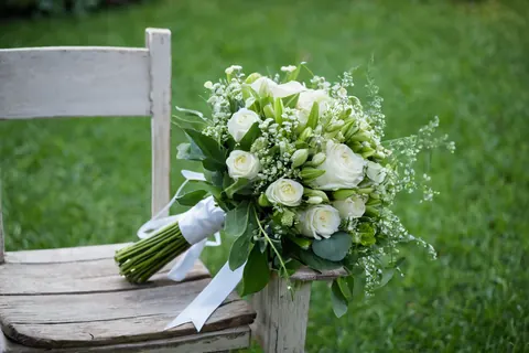 White rose bouquet with greenery tied with white ribbon on wooden chair outdoors